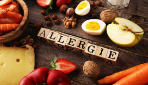 Schriftzug "Allergie" aus Holzwürfeln auf Tisch mit Lebensmitteln, die Allergien verursachen können
