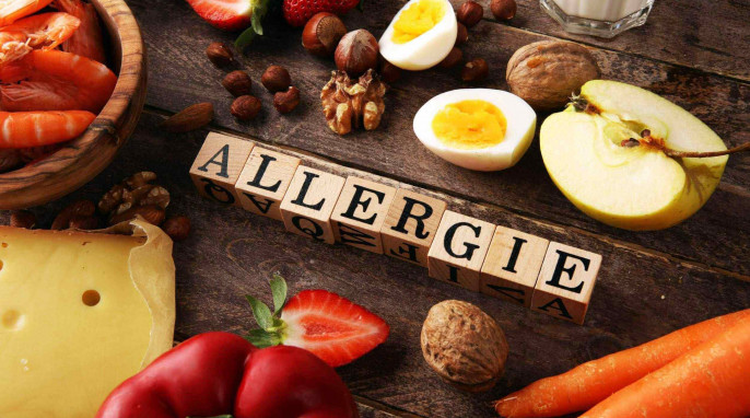 Schriftzug "Allergie" aus Holzwürfeln auf Tisch mit Lebensmitteln, die Allergien verursachen können