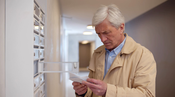 älterer Mann vor Briefkasten mit Postkarte in der Hand, schaut sorgenvoll