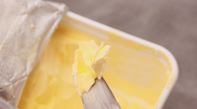 Margarine auf Messerspitze. Im Hintergrund ist eine geöffnete Maraginepackung.