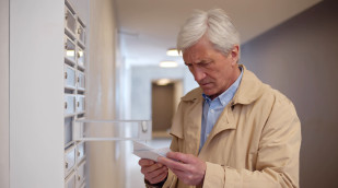 älterer Mann vor Briefkasten mit Postkarte in der Hand, schaut sorgenvoll