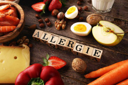 Schriftzug "Allergie" aus Holzwürfeln auf Tisch mit Lebensmitteln, die Allergien verursachen können