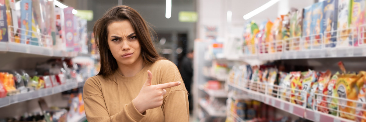 unzufriedene Verbraucherin zeigt im Supermarkt mit dem Finger auf Produkte im Regal